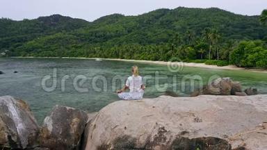 空中飞行围绕金发女孩坐在花岗岩岩石与异国情调的热带海滩和冥想。 生活方式的平衡